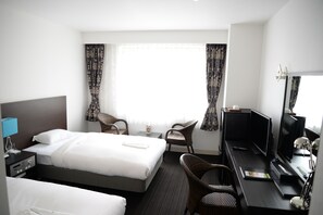 A hotel room with a bed, two chairs, a desk with a computer, a TV, and a window with curtains.