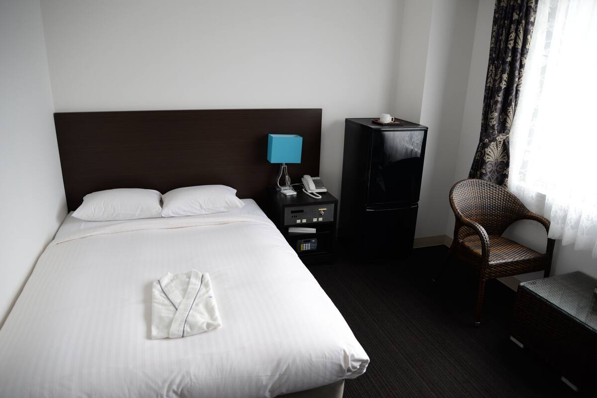 Habitación de hotel con una cama grande, una mesita de noche con lámpara, teléfono, televisor, una silla y una pequeña mesa.