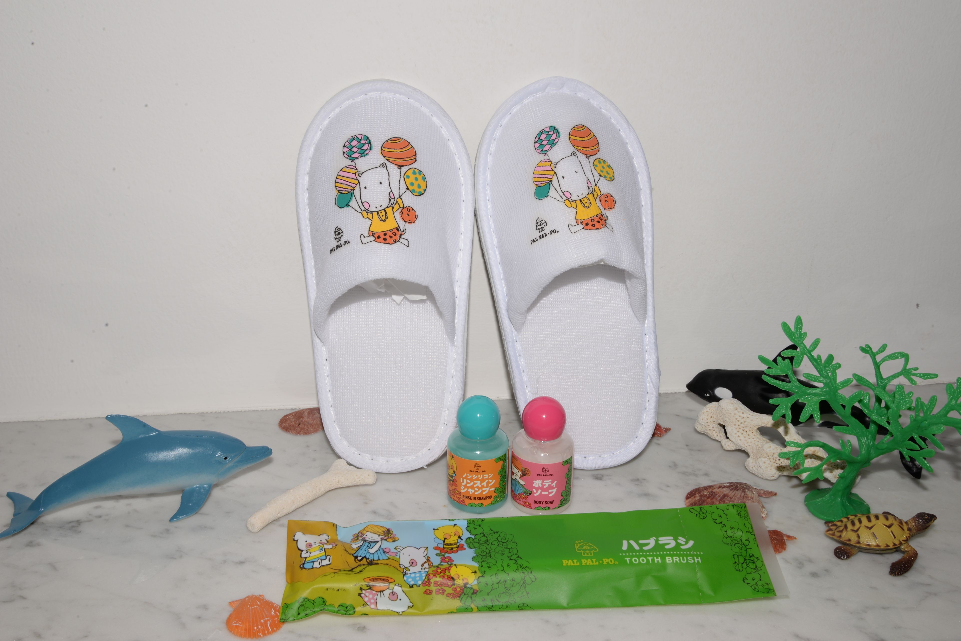 Ein Paar weiße Hausschuhe mit Zeichentrickfiguren, ein Dophin-Spielzeug, eine Zahnbürste und eine grüne Zahnbürstenverpackung.