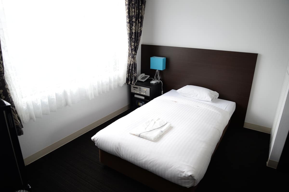 Una habitación de hotel con una cama bien hecha, una mesita de noche con lámpara y teléfono, y un ventanal con cortinas translúcidas.