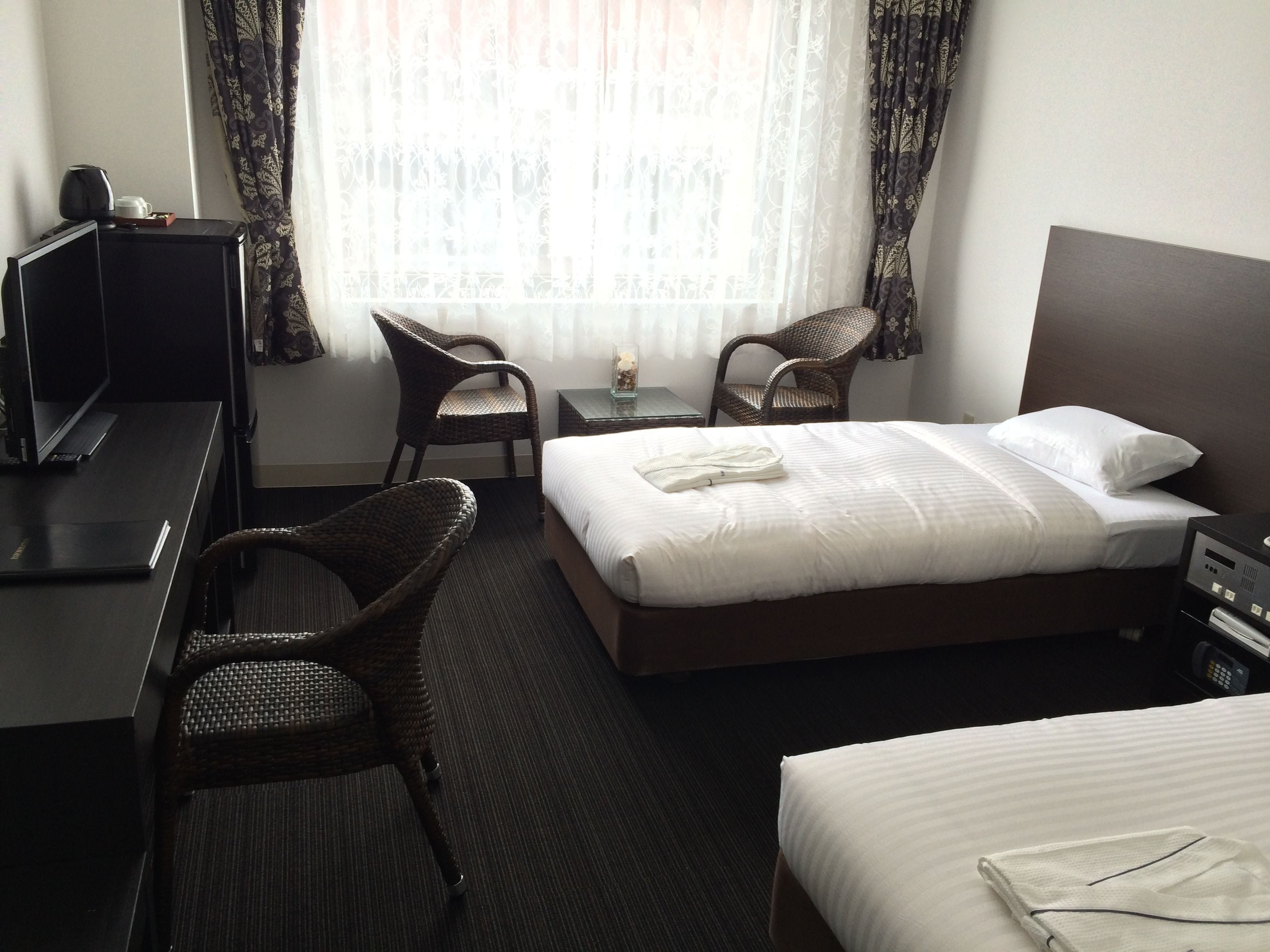 Une chambre d’hôtel avec un lit, deux fauteuils en osier, un bureau avec un ordinateur et une petite table avec une lampe.