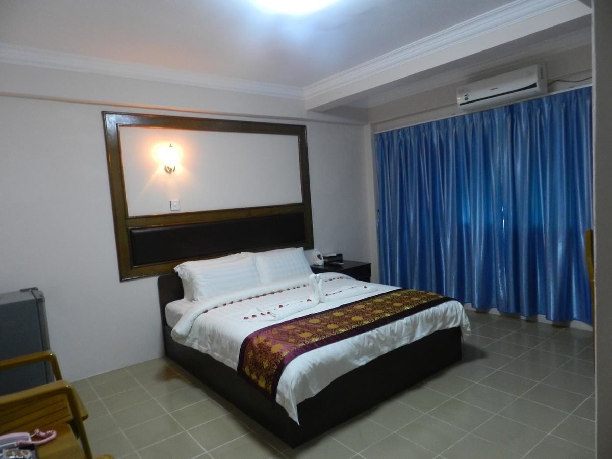 A hotel room with a bed, a nightstand, a wall-mounted light, and a window with blue curtains.