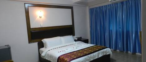 A hotel room with a bed, a nightstand, a wall-mounted light, and a window with blue curtains.