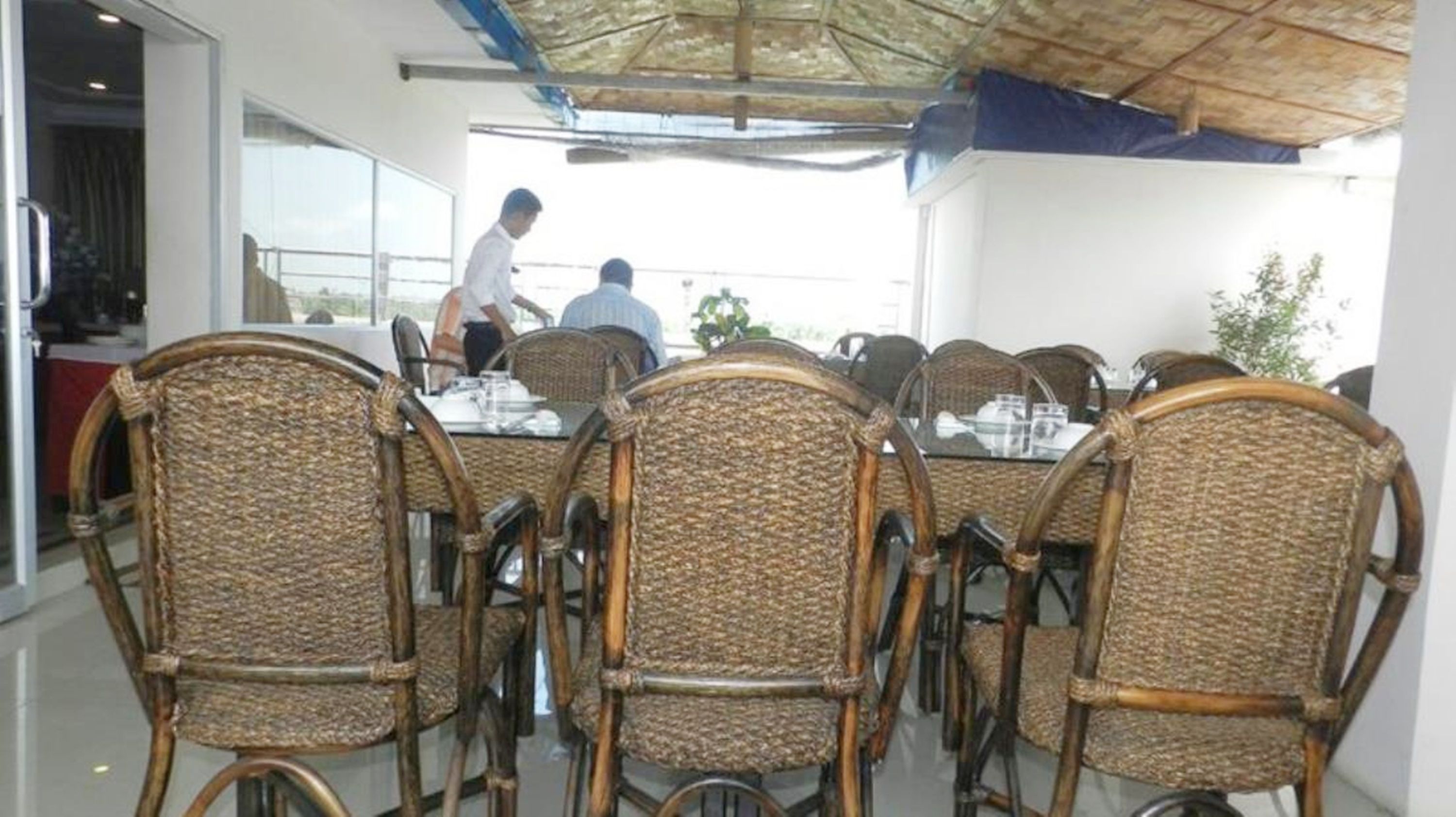 A dining area with wicker chairs and a glass table, a person walking in the background, and a view of the outdoors.