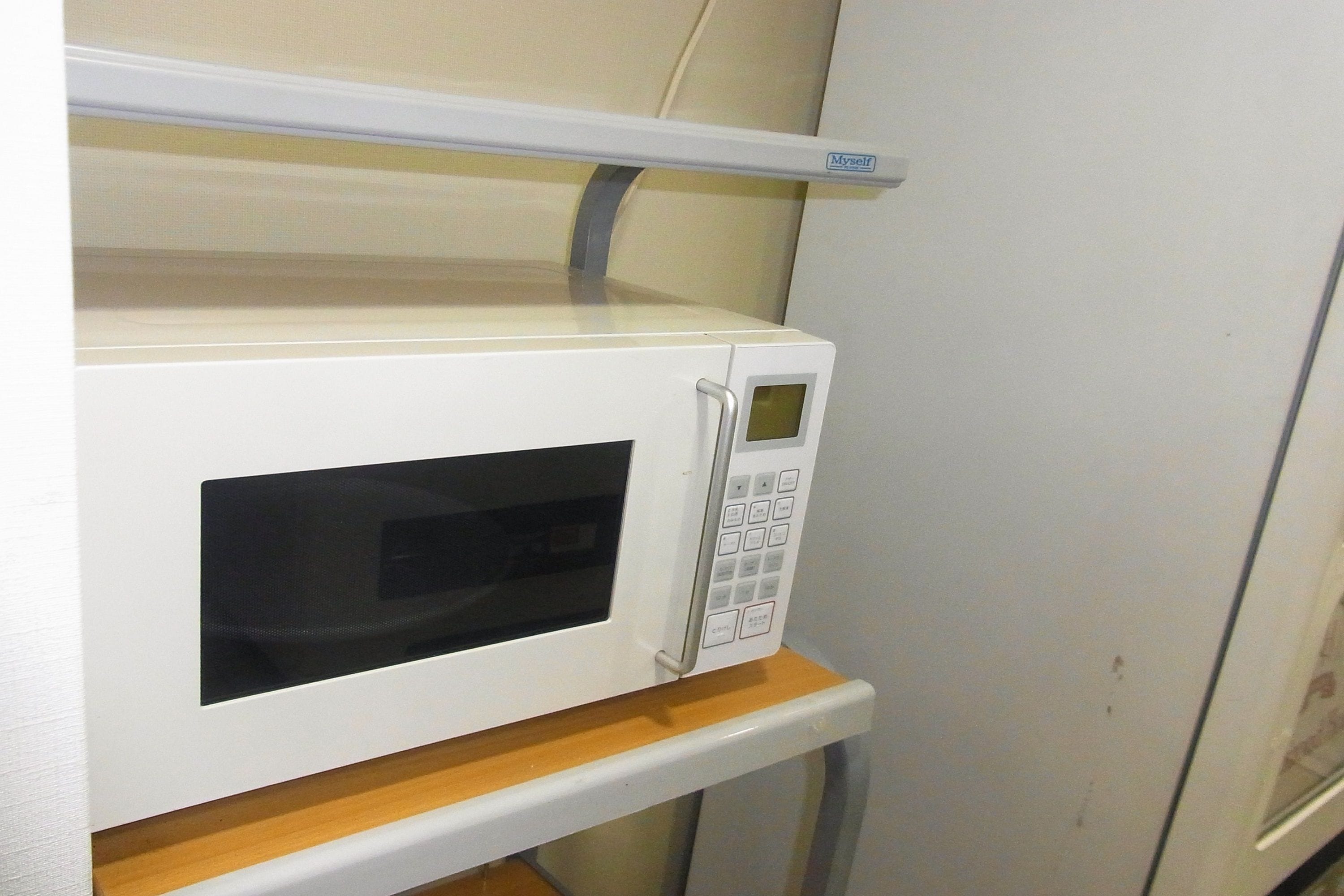 A white microwave oven with a digital display and control panel, mounted on a wooden shelf above a white refrigerator.