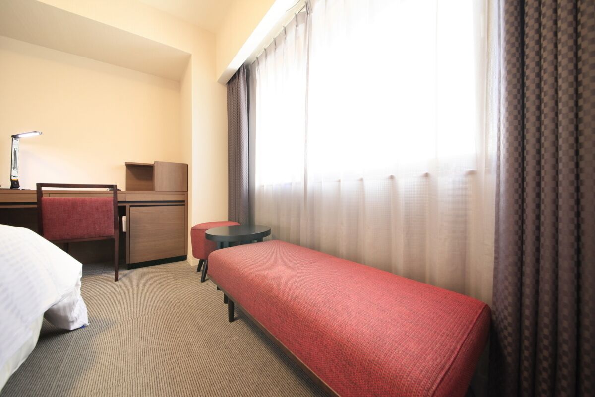 A hotel room with a bed, a desk with a chair, a small table, and a window with curtains.