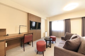 Ein Hotelzimmer mit Schreibtisch, Fernseher, Sofa und Pouf.