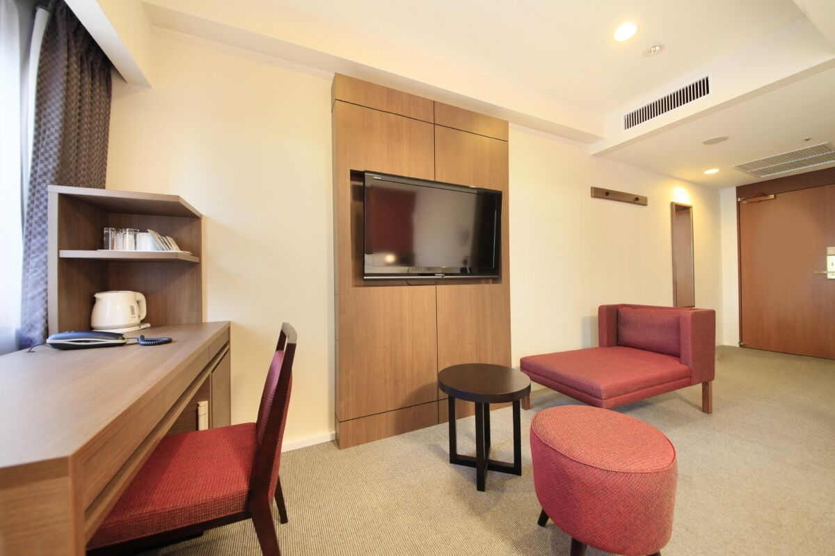 A hotel room with a wooden desk, a television mounted on a cabinet, a red chair, a small round table, and a red ottoman.