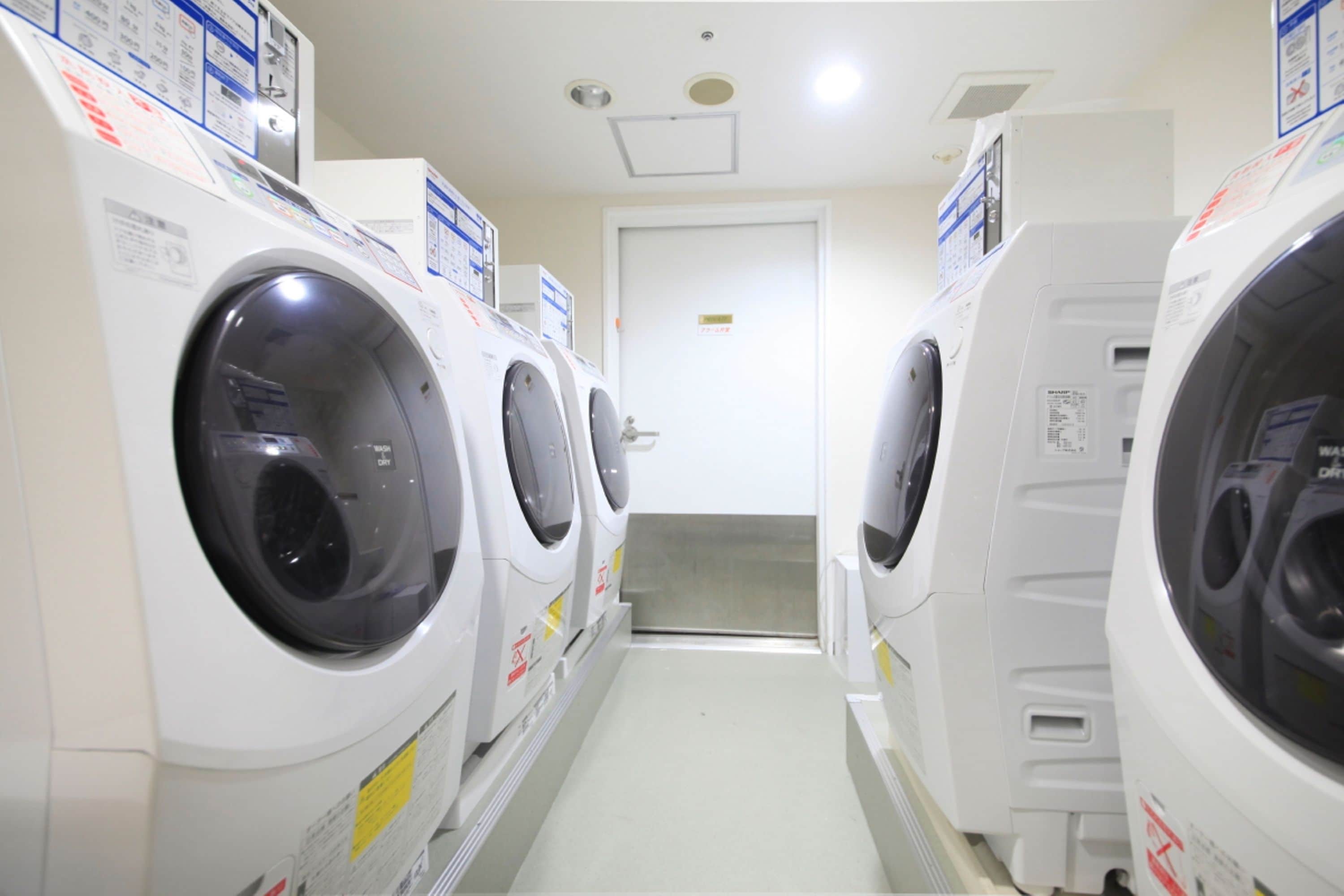 A laundry room with multiple washing machines lined up against the wall, a door at the end, and a tiled floor.