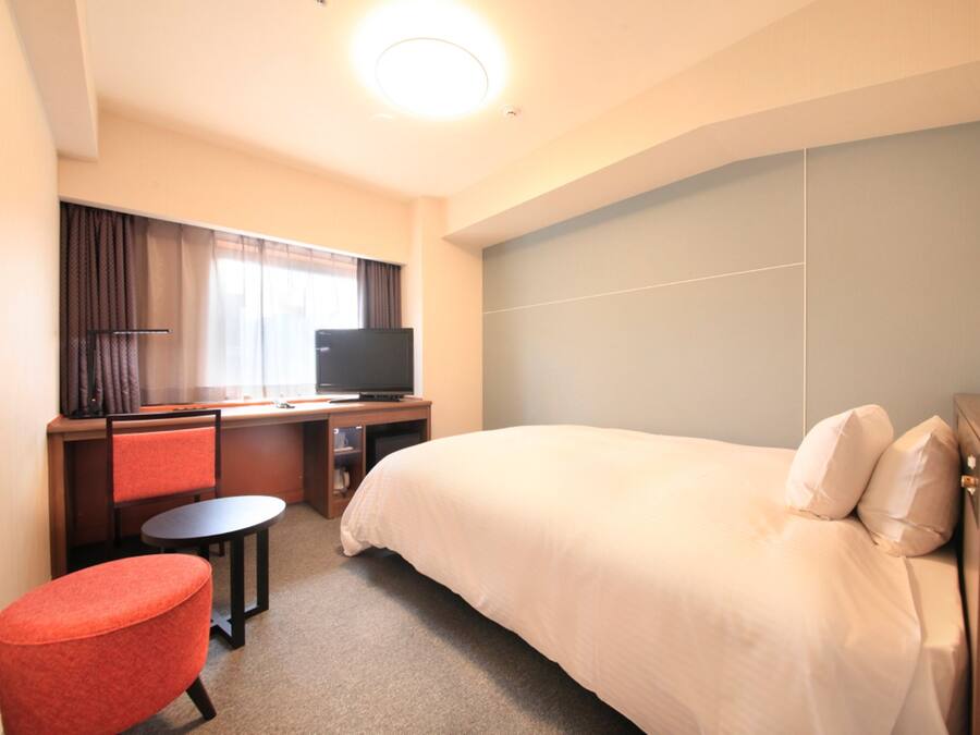 Una habitación de hotel con cama, escritorio con televisor, una mesita y una silla roja.