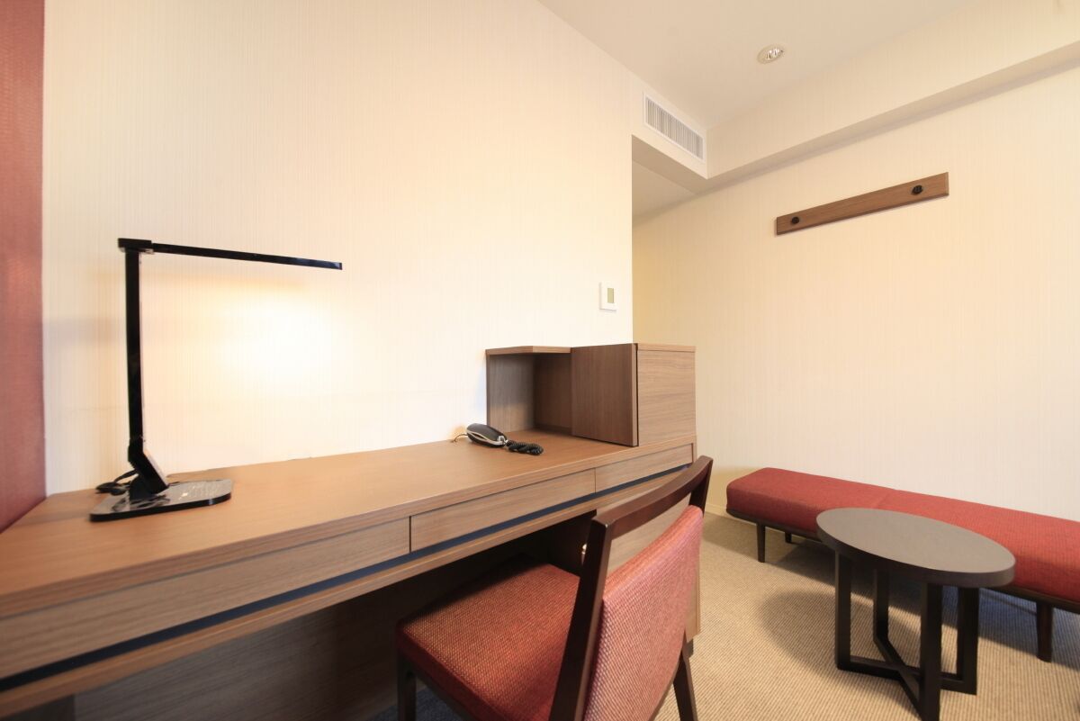 A hotel room with a wooden desk, a chair, a small round table, a red bench, and a wall-mounted shelf.