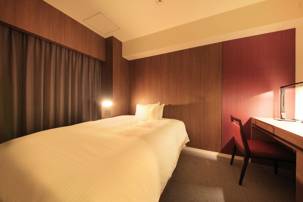 A hotel room with a large bed, a desk with a chair, a wall-mounted lamp, and a window with curtains.