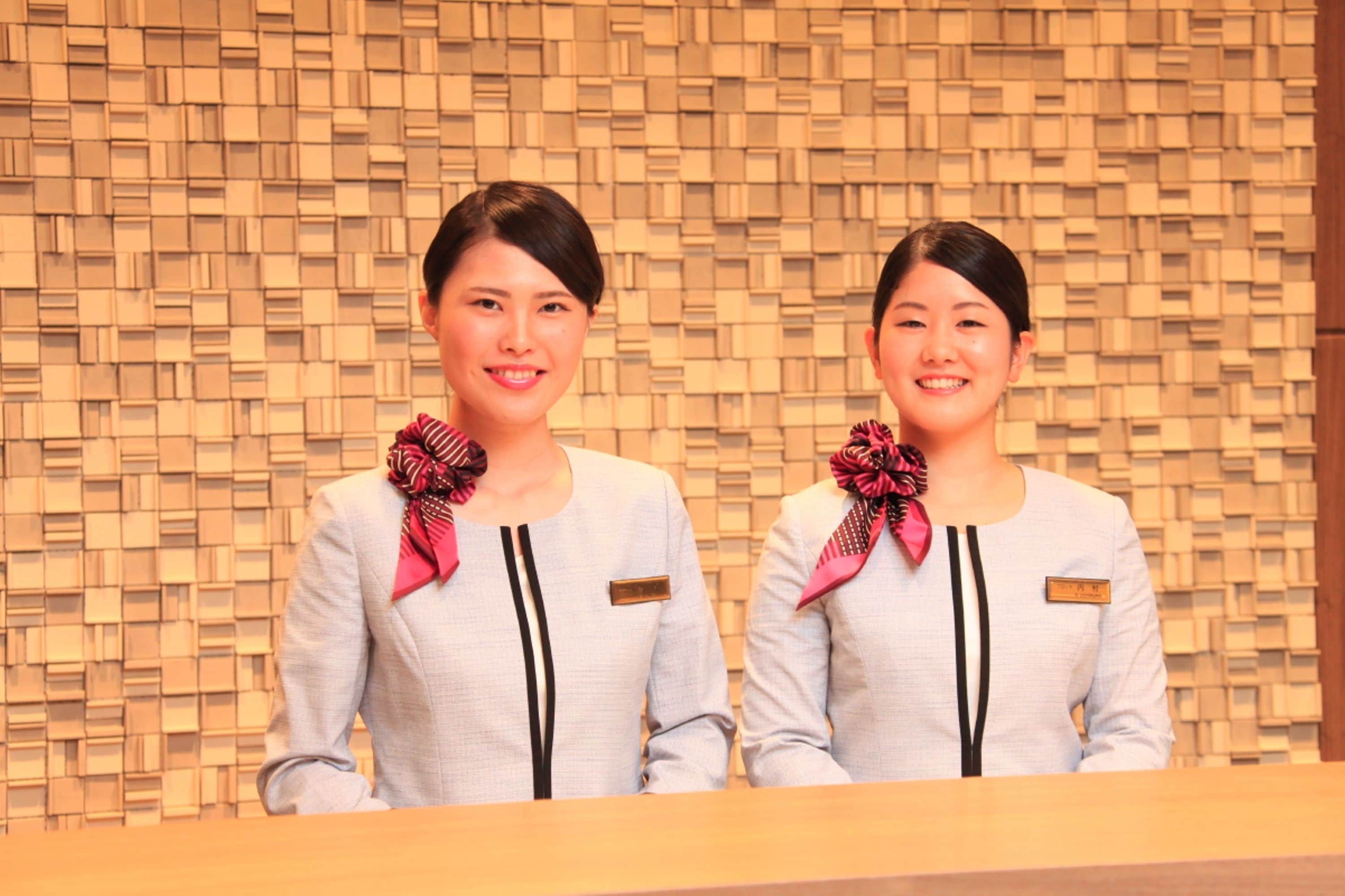 Two receptionists standing in front of a patterned wall, wearing light grey uniforms with name tags.
