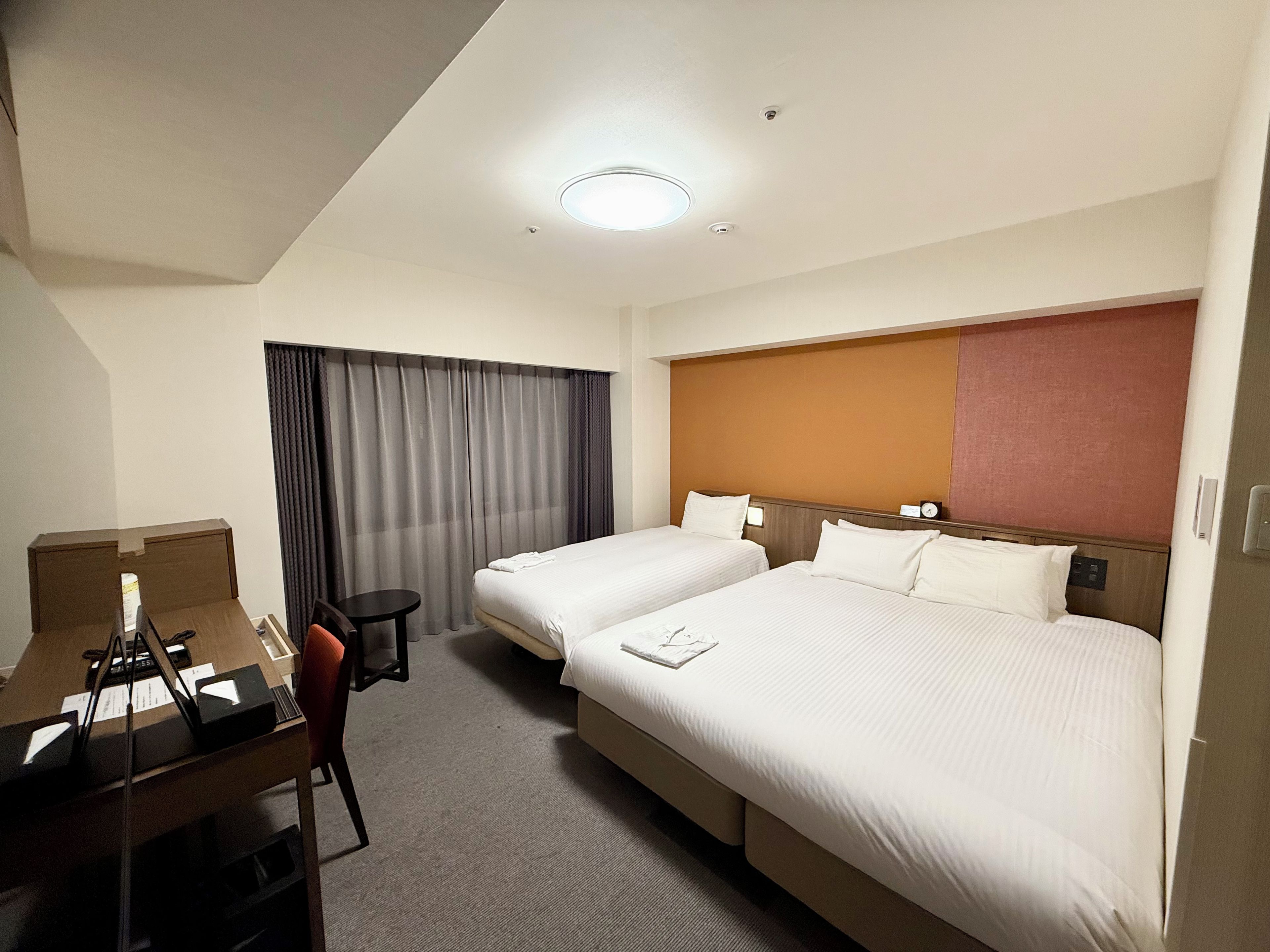 Habitación de hotel con dos camas, un escritorio, una silla, una mesita y un ventanal con cortinas.