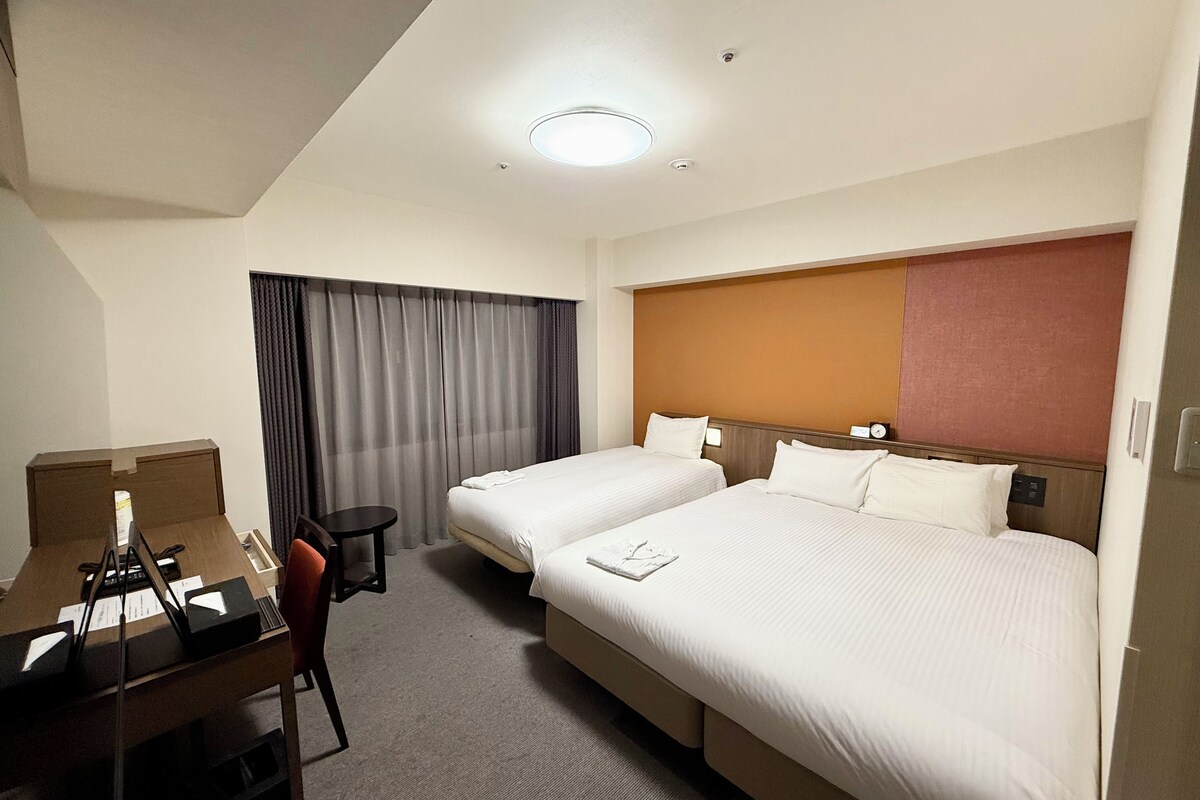 A hotel room with two beds, a desk, a chair, a small table, and a window with curtains.