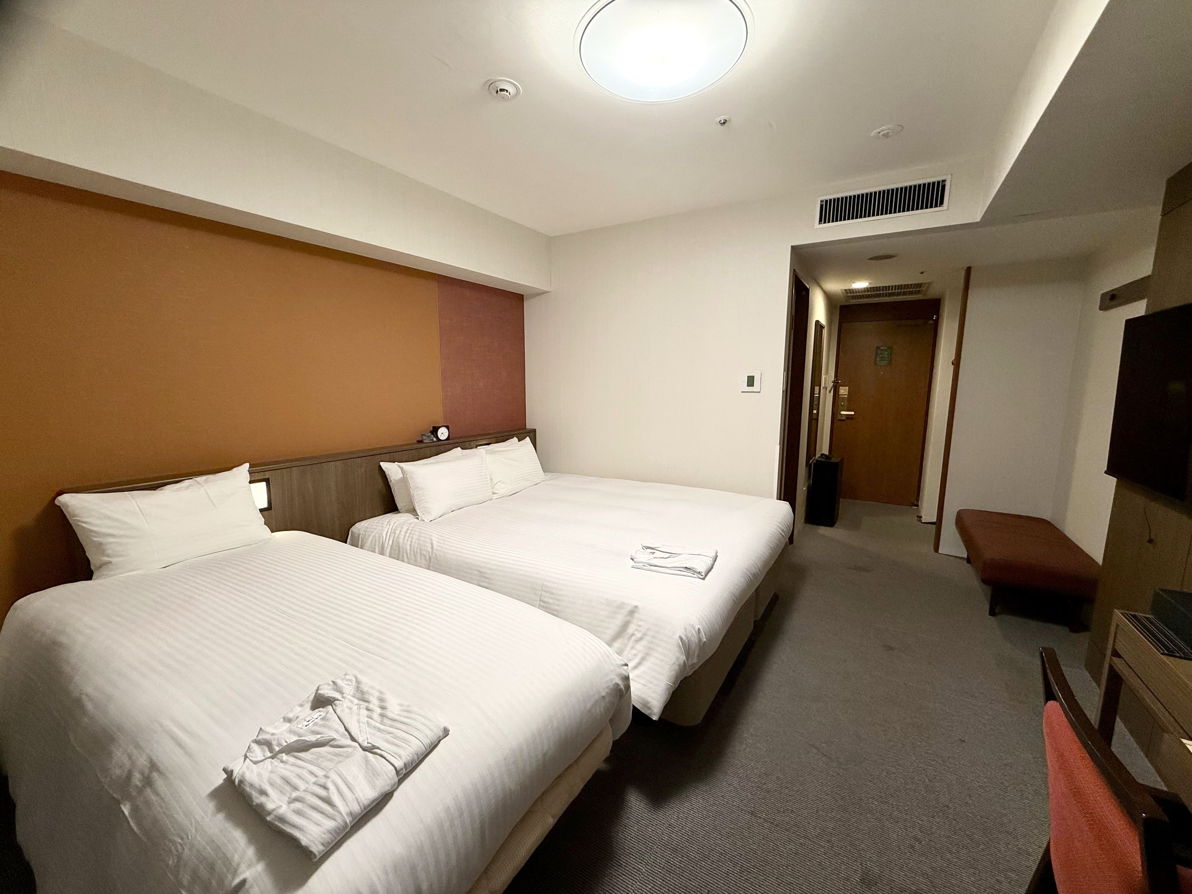 Ett hotellrum med två sängar, en platt-TV, en bänk och en dörr som leder till ett annat rum.