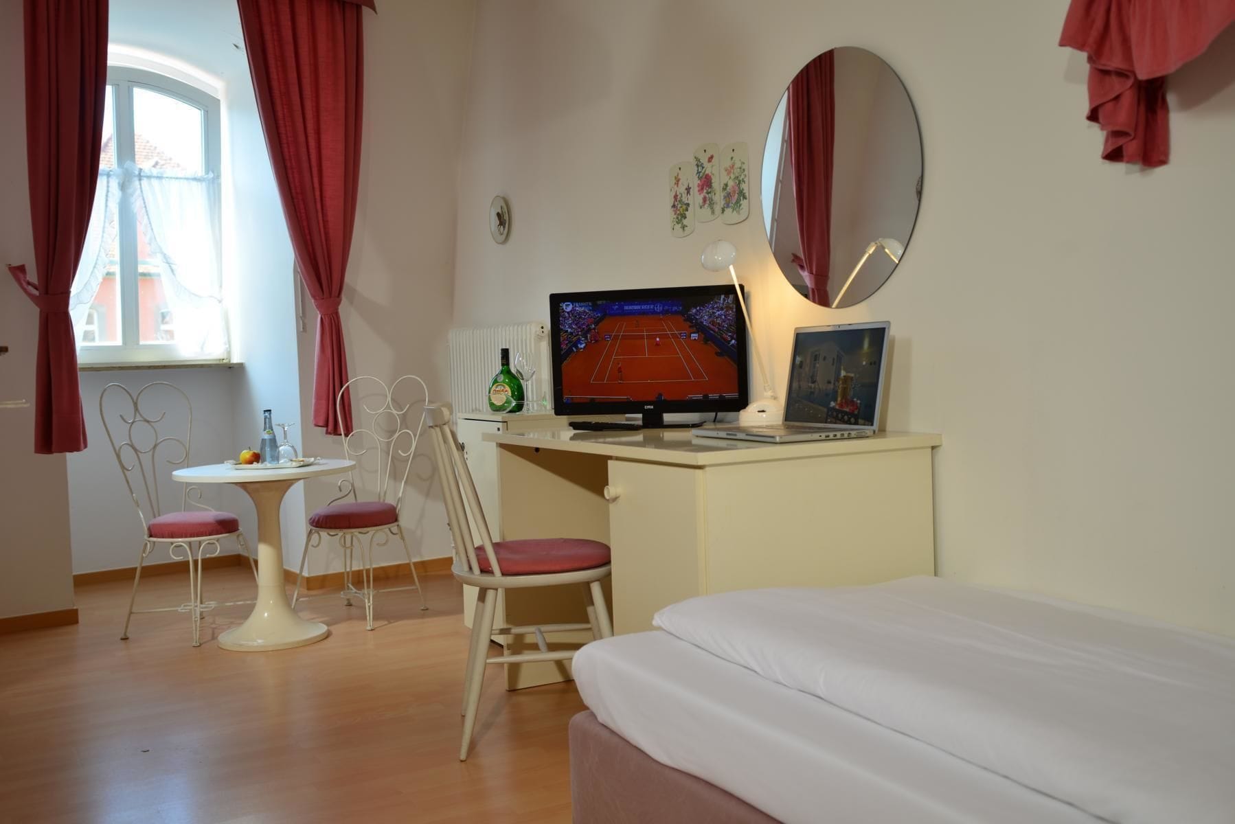 Ein Zimmer mit einem Bett, einem Schreibtisch mit Computer, zwei Stühlen, einem Fenster mit Vorhängen und einem Spiegel an der Wand.