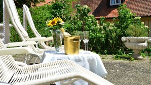 Een tuinsetting met witte lounge fauteuils, een tafel met een witte tafelkleed, een champagnefles en twee wijnglazen.