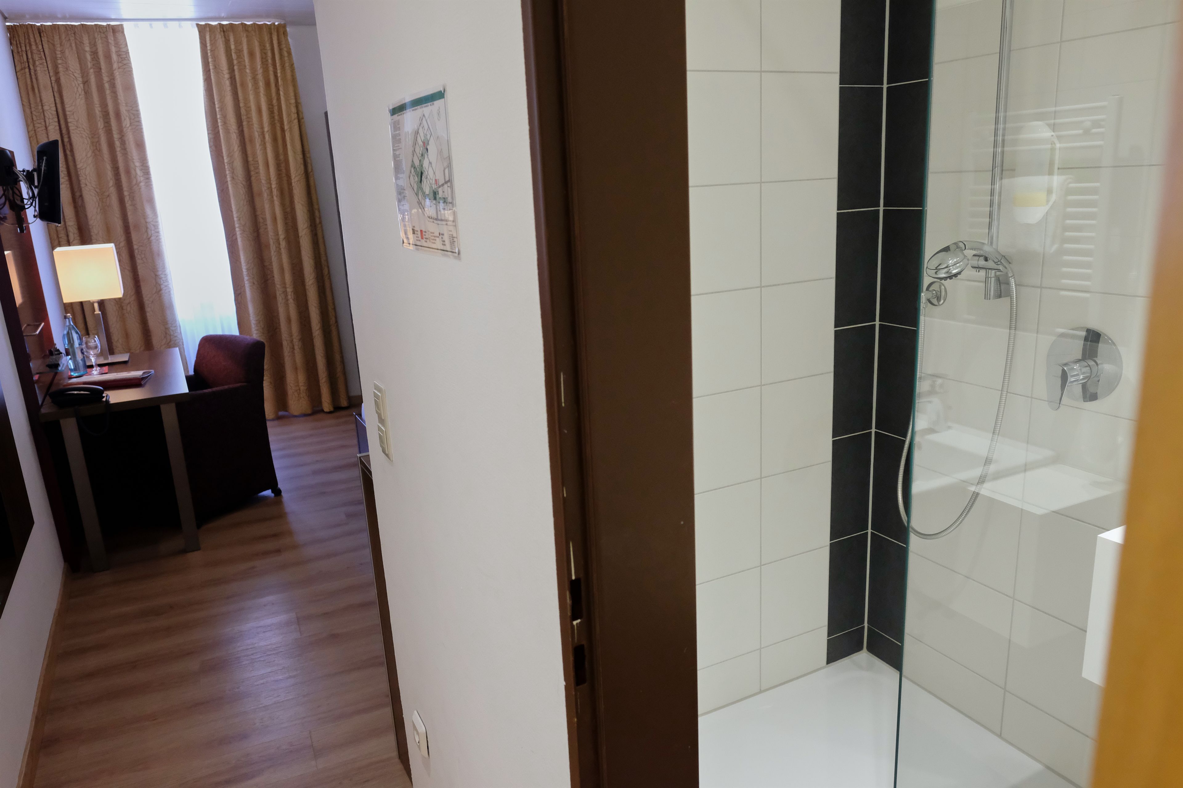 Habitación de hotel con piso de madera, escritorio con lámpara, una silla y zona de ducha con puerta de vidrio.