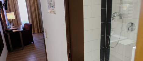 Habitación de hotel con piso de madera, escritorio con lámpara, una silla y zona de ducha con puerta de vidrio.