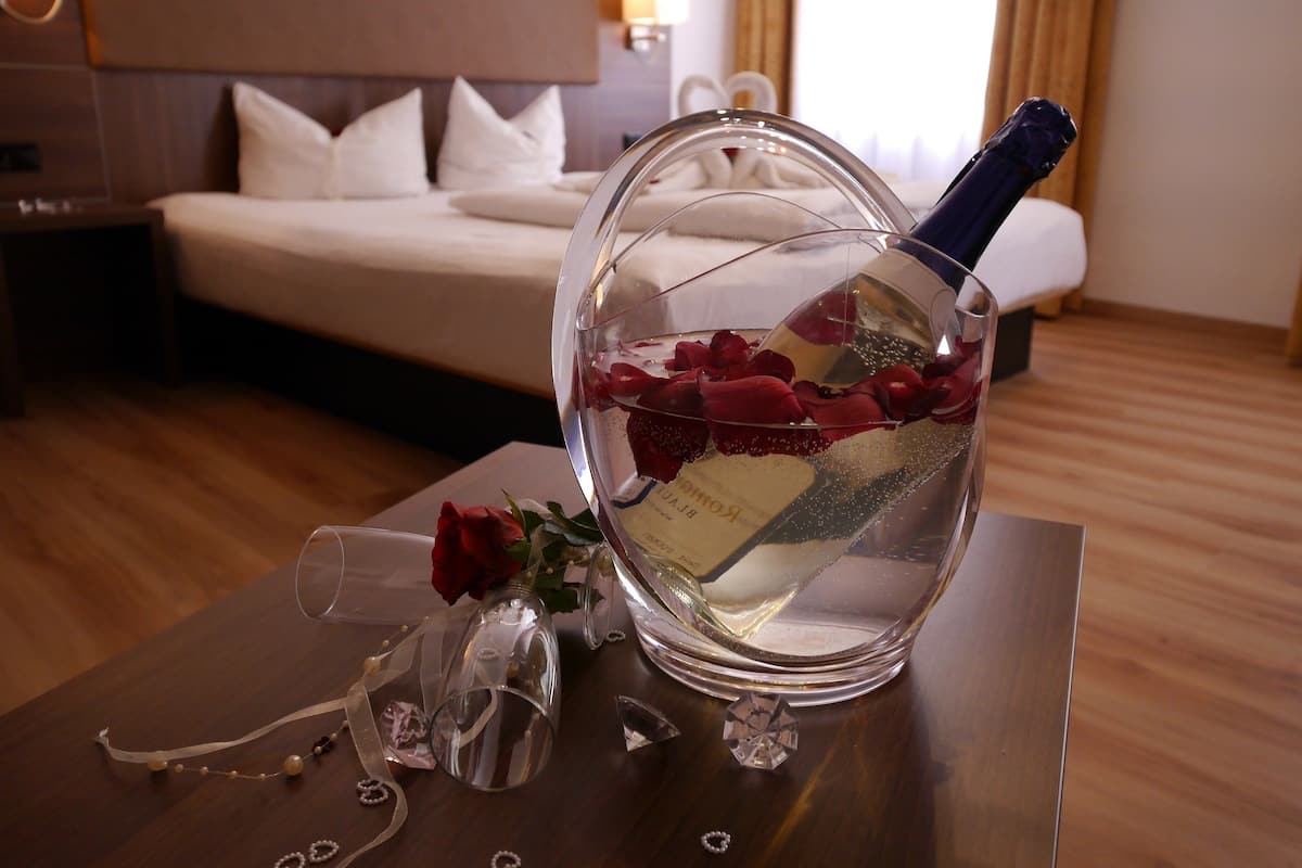 Ein Hotelzimmer mit einem Bett, einem Nachttisch mit Champagner, Rosenblättern und einem dekorativen Glas.
