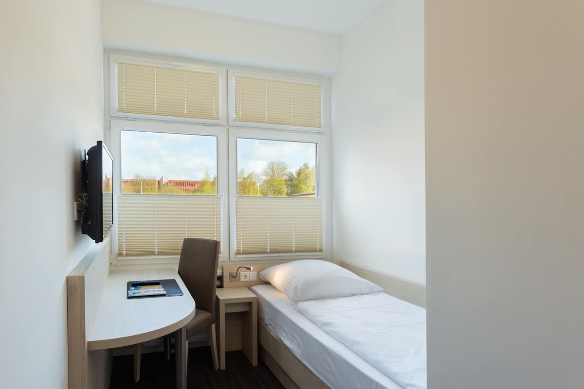 Quarto pequeno com uma cama, uma escrivaninha e uma cadeira. Há duas janelas com persianas e uma televisão presa à parede.