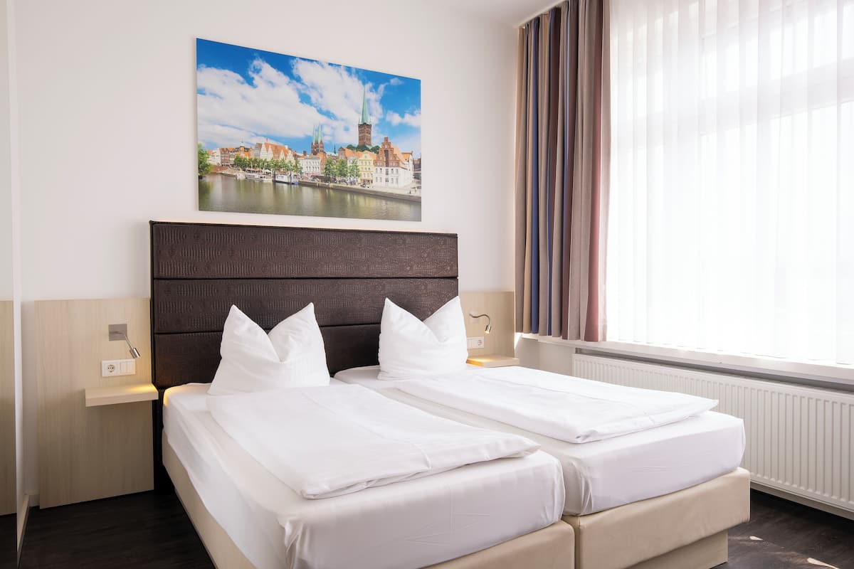 Quarto de hotel com cama grande, cabeceira, mesa de cabeceira, quadro pendurado na parede e janela com cortinas.