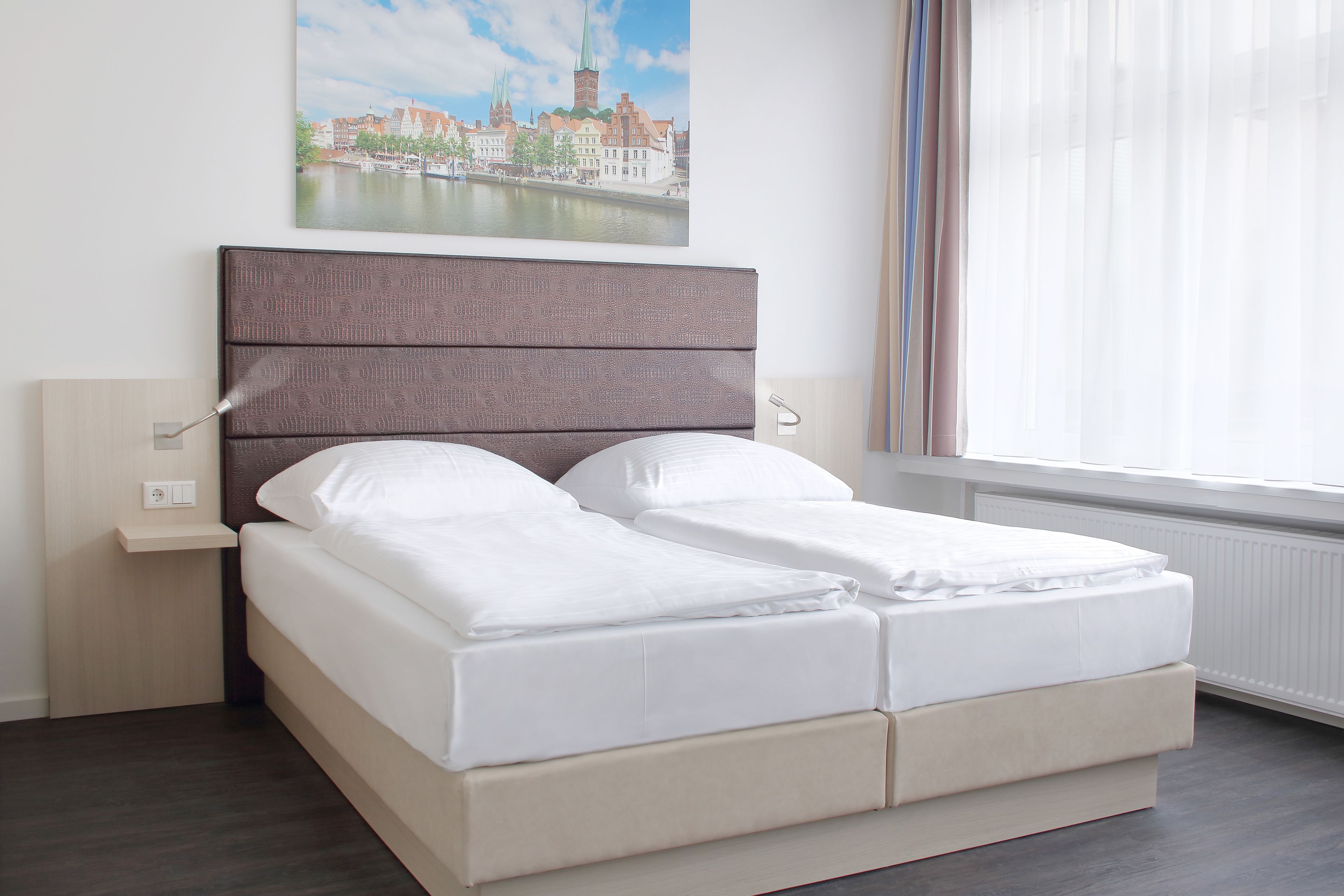 Ein Doppelbett mit Kopfteil, Nachttisch und einem Fenster mit durchsichtigen Vorhängen.