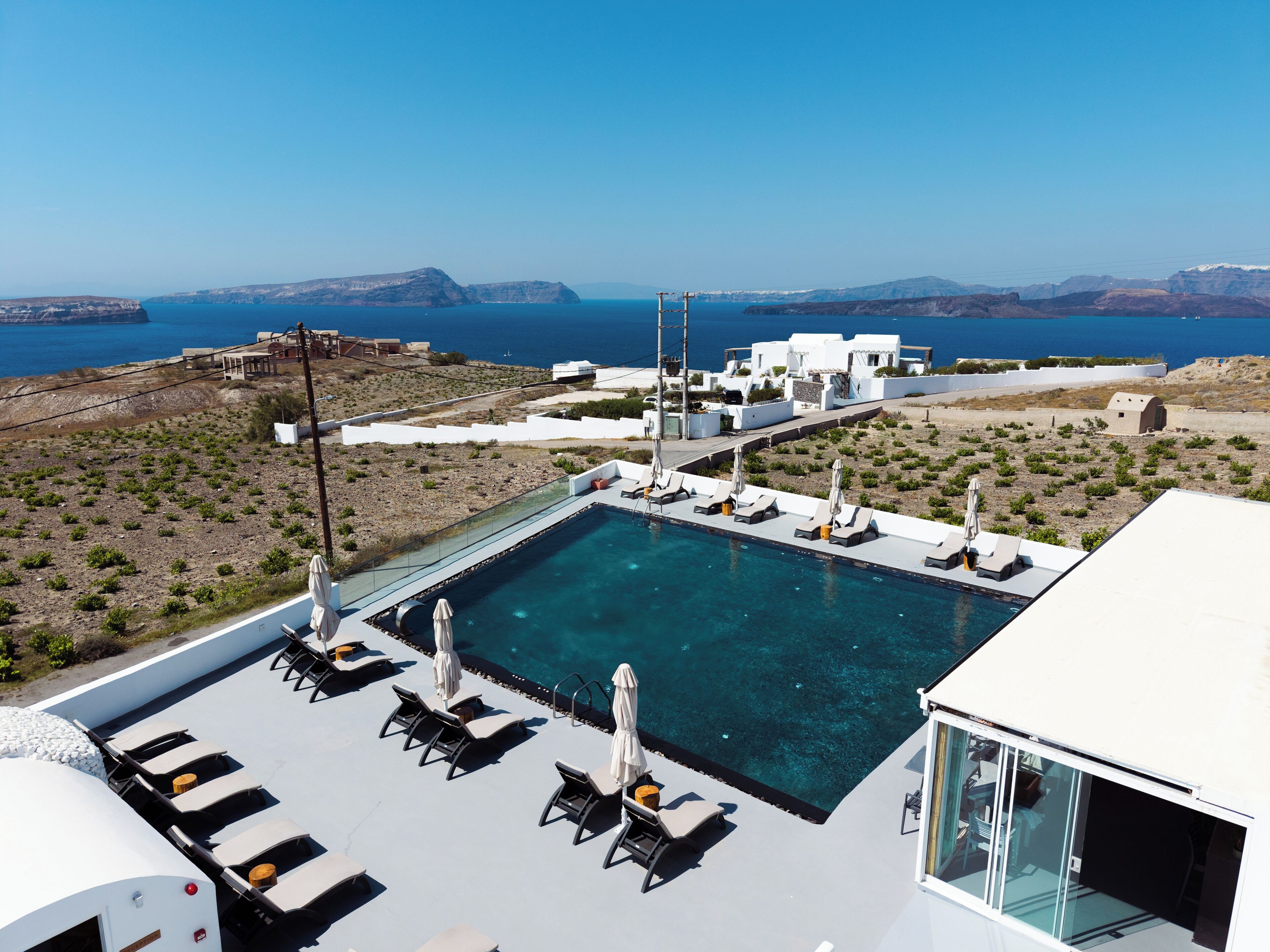 A rooftop infinity pool with lounge chairs and umbrellas, overlooking a coastal landscape with buildings and a clear blue sky.