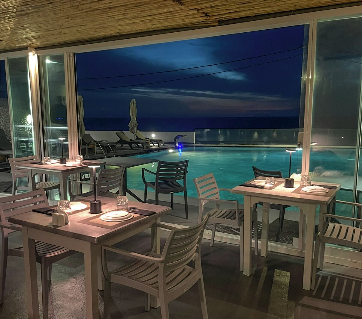 An outdoor dining area with tables and chairs, overlooking a pool and the ocean at night.