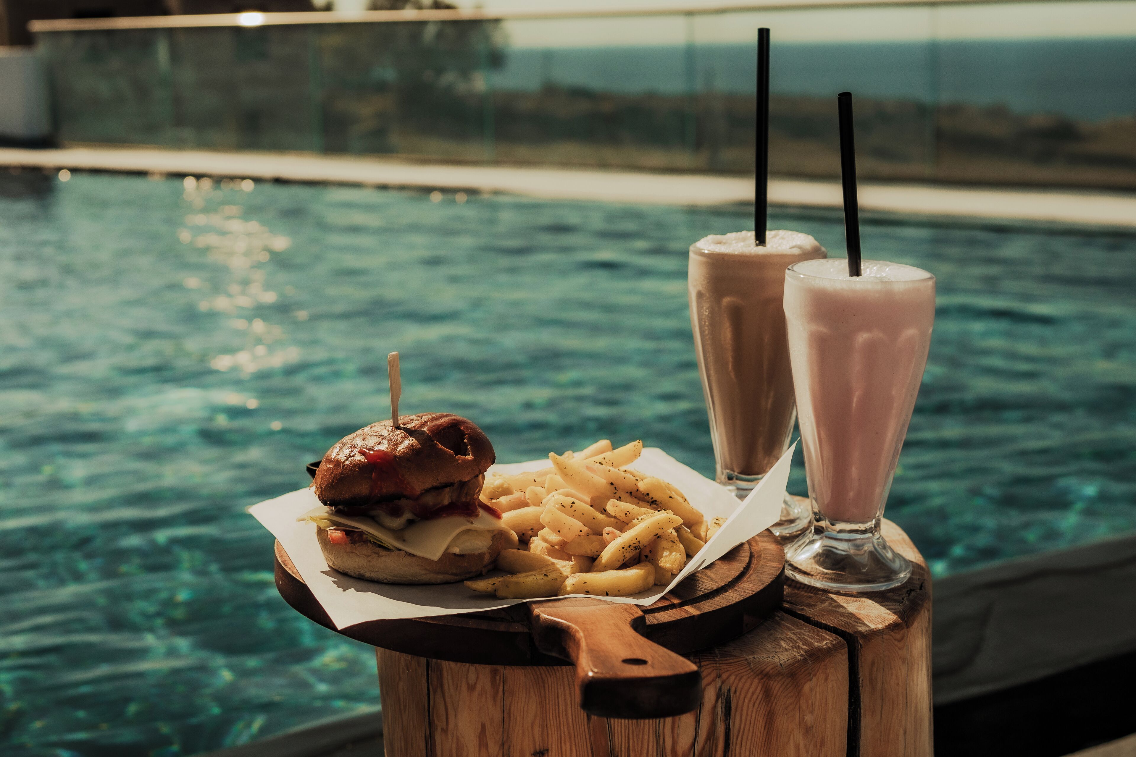 Un hamburger con patatine su un tagliere di legno, accompagnato da due frappè, posto vicino a una piscina.