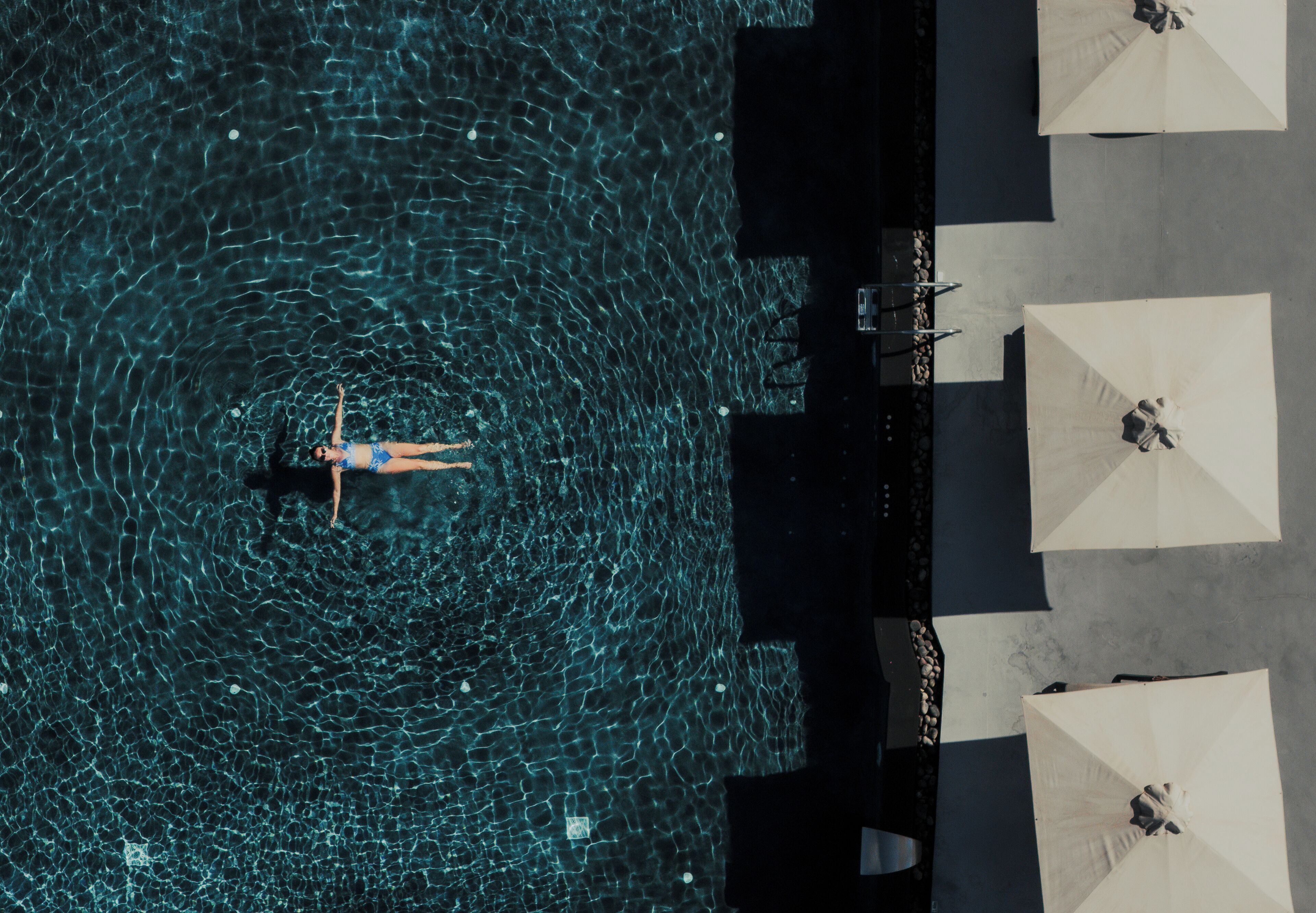 Vista aérea de uma piscina com alguém nadando, cercada por guarda-sóis e espreguiçadeiras.
