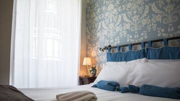 Una cama bien hecha con ropa de cama azul y blanca, una lámpara en una mesita de noche y una ventana con cortinas traslúcidas.