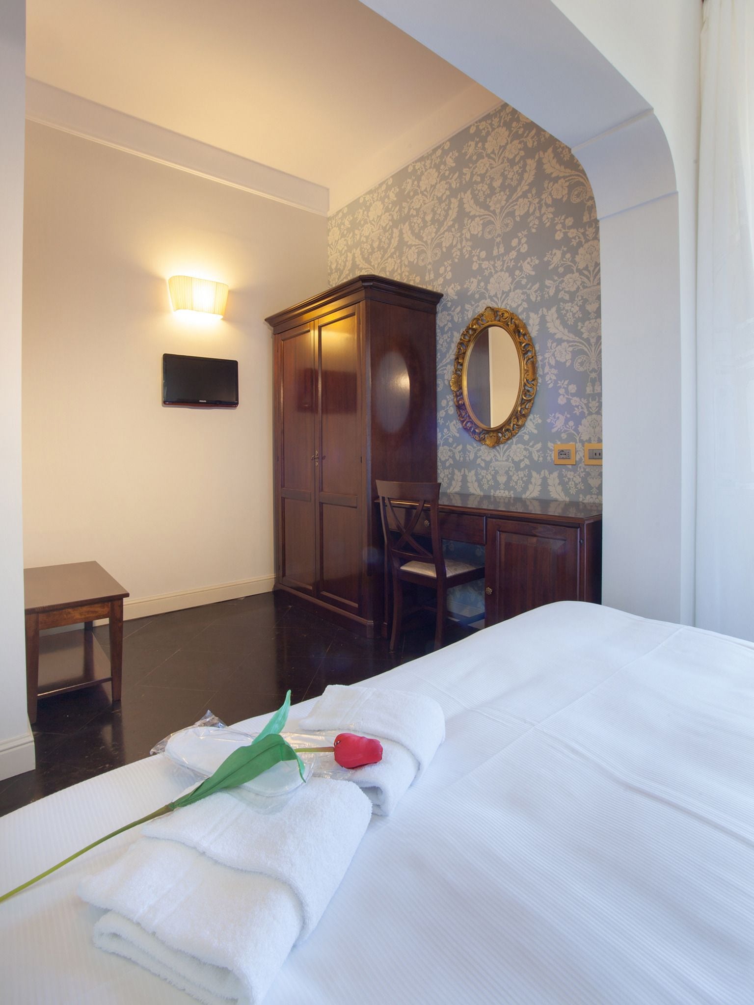 A hotel room with a bed, a wardrobe, a desk, a chair, a wall-mounted light, and a mirror.