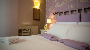 Un lit bien fait, avec du linge de lit blanc et des coussins violets, une petite table de chevet avec une lampe, et une chaise dans une pièce dont le papier peint est à motifs et qui est dotée d’un luminaire fixé au mur.