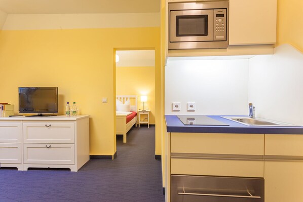 Una habitación de hotel compacta con kitchenette, una cama con manta roja, un televisor y un escritorio con cajones.