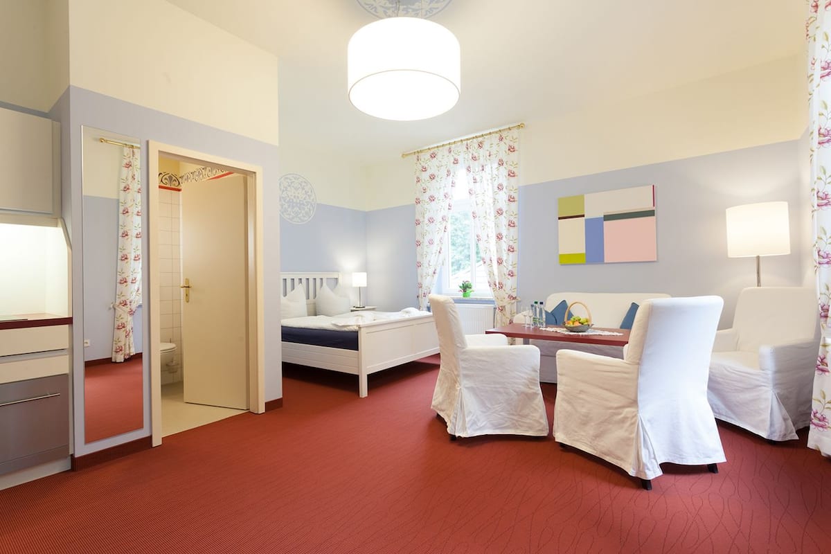 A hotel room with a bed, a desk, two chairs, a table, a window with floral curtains, a bedside lamp, and a wall clock.