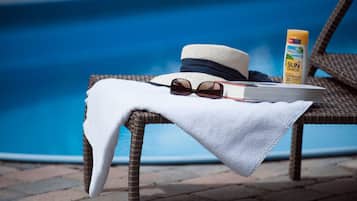 A wicker lounge chair by a pool with a white towel, a hat, sunglasses, and a bottle of sunscreen.