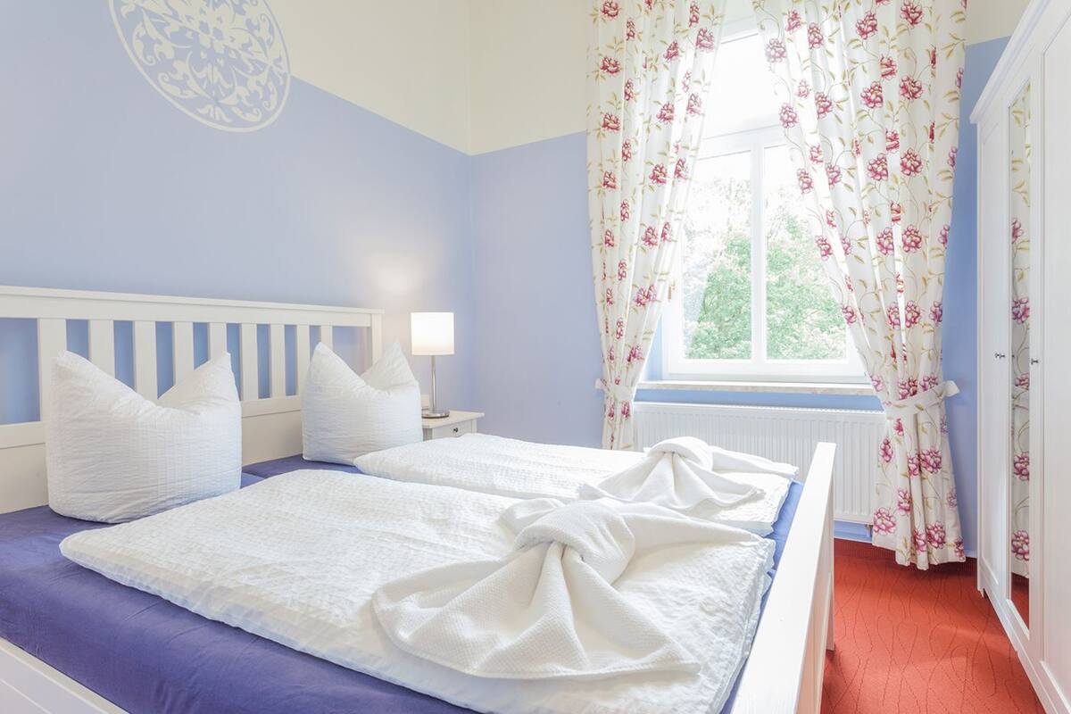 A bedroom with two beds, a window with floral curtains, and a white headboard.