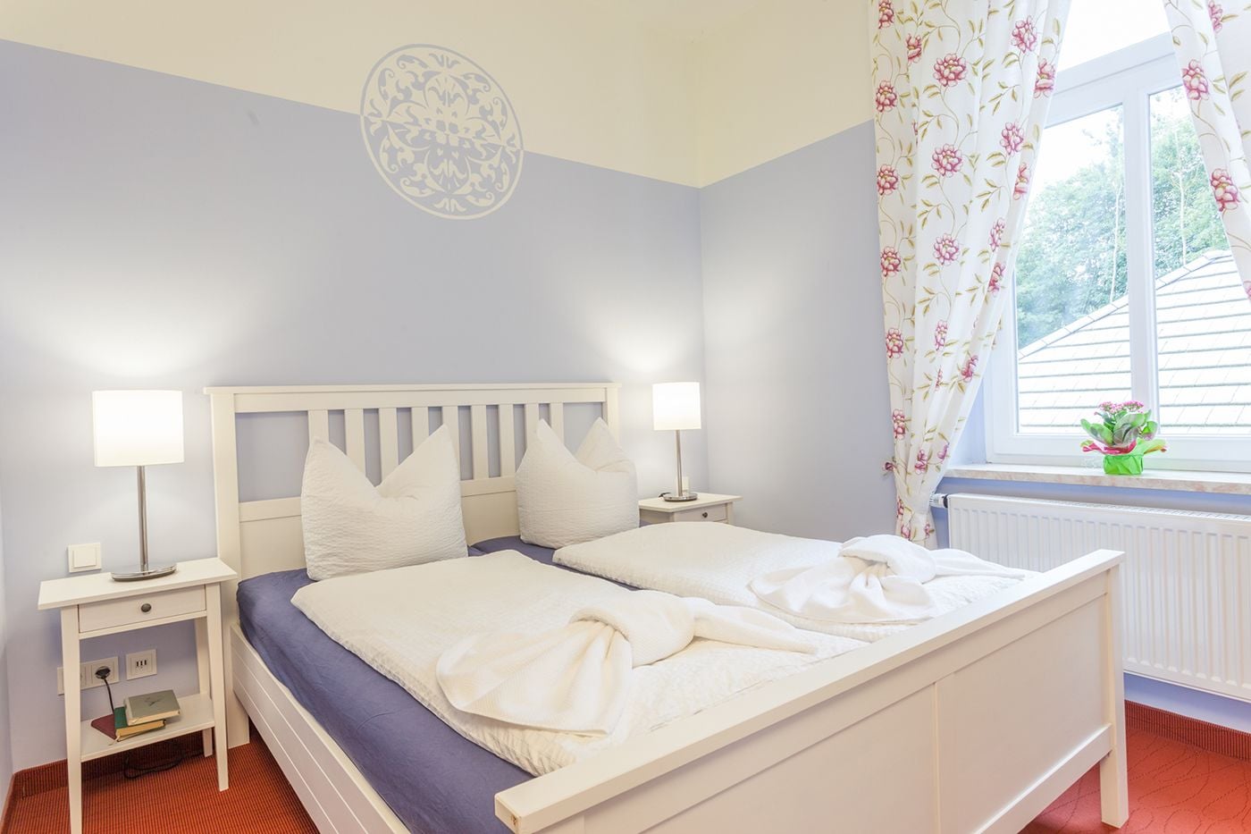 A bedroom with a white bed, two bedside tables with lamps, a window with floral curtains, and a radiator.