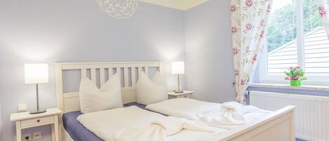 A bedroom with a white bed, two bedside tables with lamps, a window with floral curtains, and a radiator.