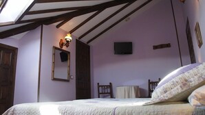 A bedroom with a sloped ceiling, a bed with a floral bedspread, a wooden chair, a small round table, and a mounted television. - Posada La Cerrá de San Roque (Santillana del Mar)