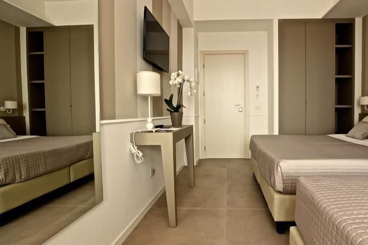 A hotel room with two beds, a TV mounted on the wall, a desk with a lamp, and a door leading to another room.