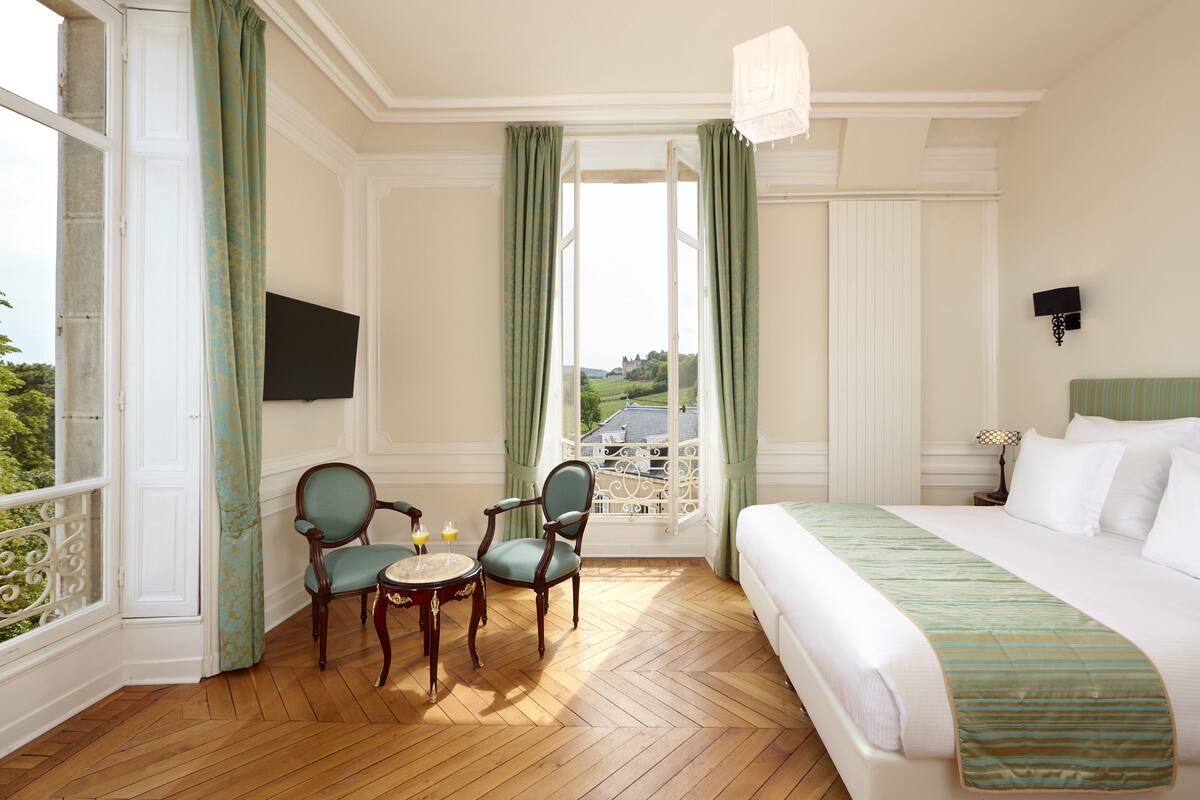 Habitació d'hotel amb un llit, dues cadires, una taula petita amb gots, un televisor a la paret i vista d'un castell per la finestra.