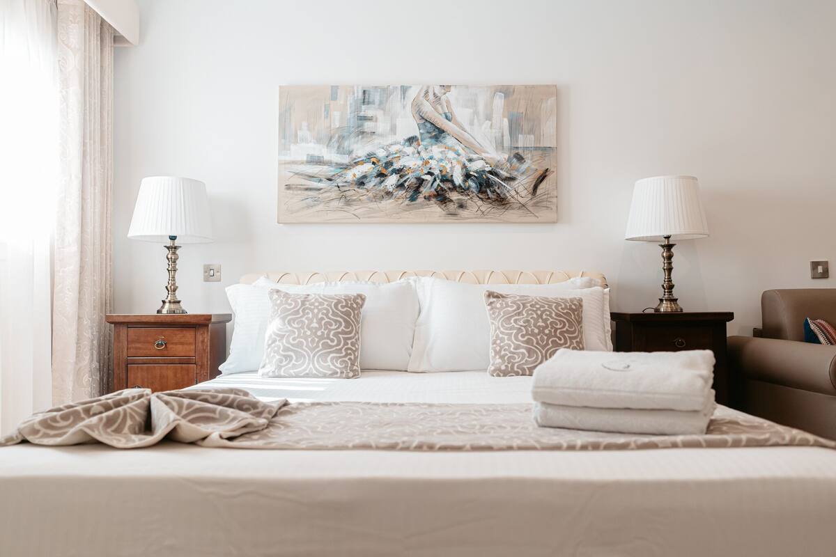 Una cama bien hecha con almohadones estampados, una mesita de noche con lámpara, y un cuadro abstracto de gran tamaño en la pared.