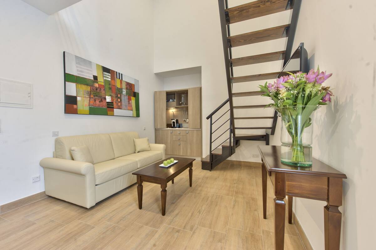 Una sala de estar con un sofá, una mesa de centro y un florero. Se ve una escalera al fondo.