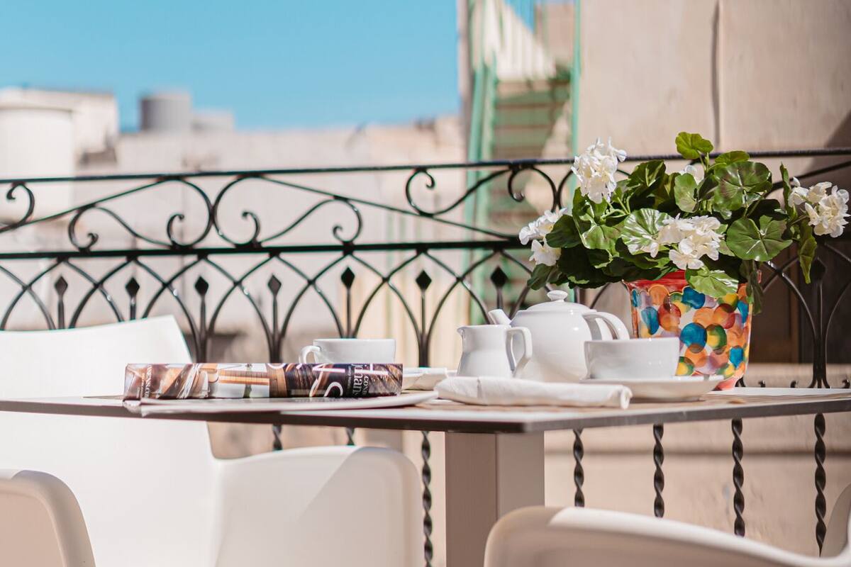 Un balcón con una mesa dispuesta para el té, sillas blancas y una maceta de flores coloridas.