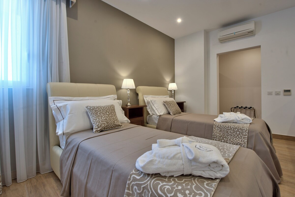 Una habitación de hotel con dos camas, lámparas de noche, un equipo de aire acondicionado y una ventana con cortinas traslúcidas.