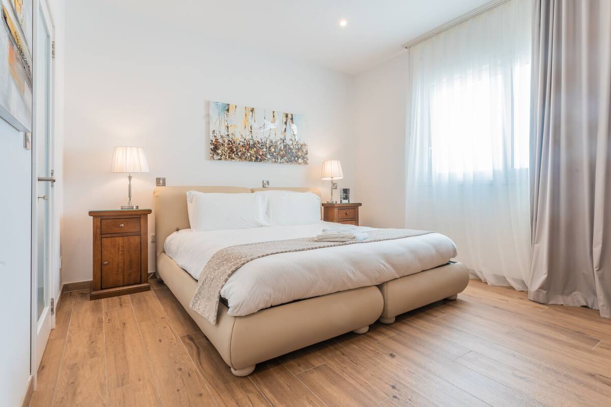 Un dormitorio con una cama grande, mesitas de noche de madera, un cuadro en la pared y una ventana con cortinas.