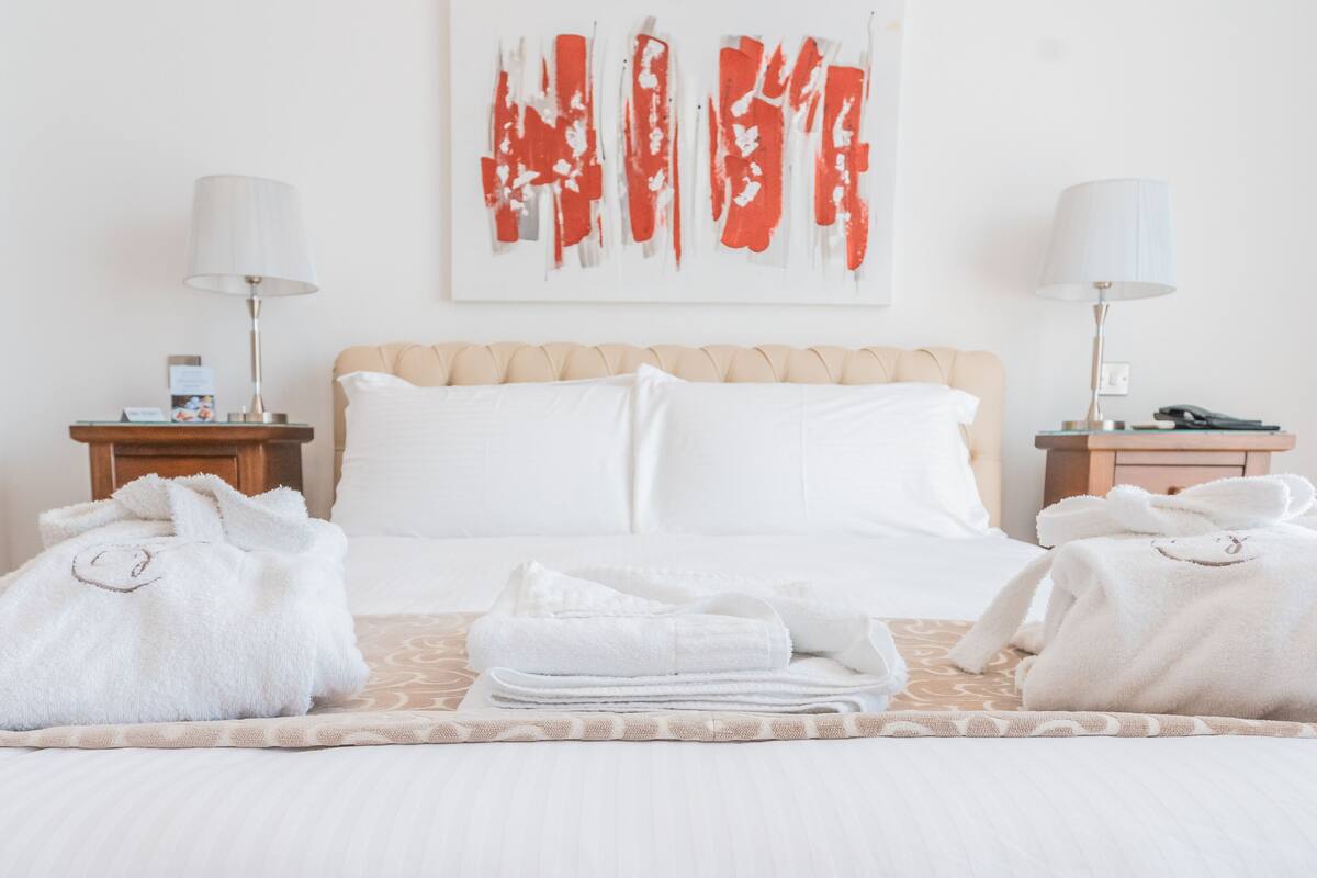 Una cama bien hecha con sábanas y toallas blancas, un cabecero color beige y un cuadro abstracto rojo en la pared.